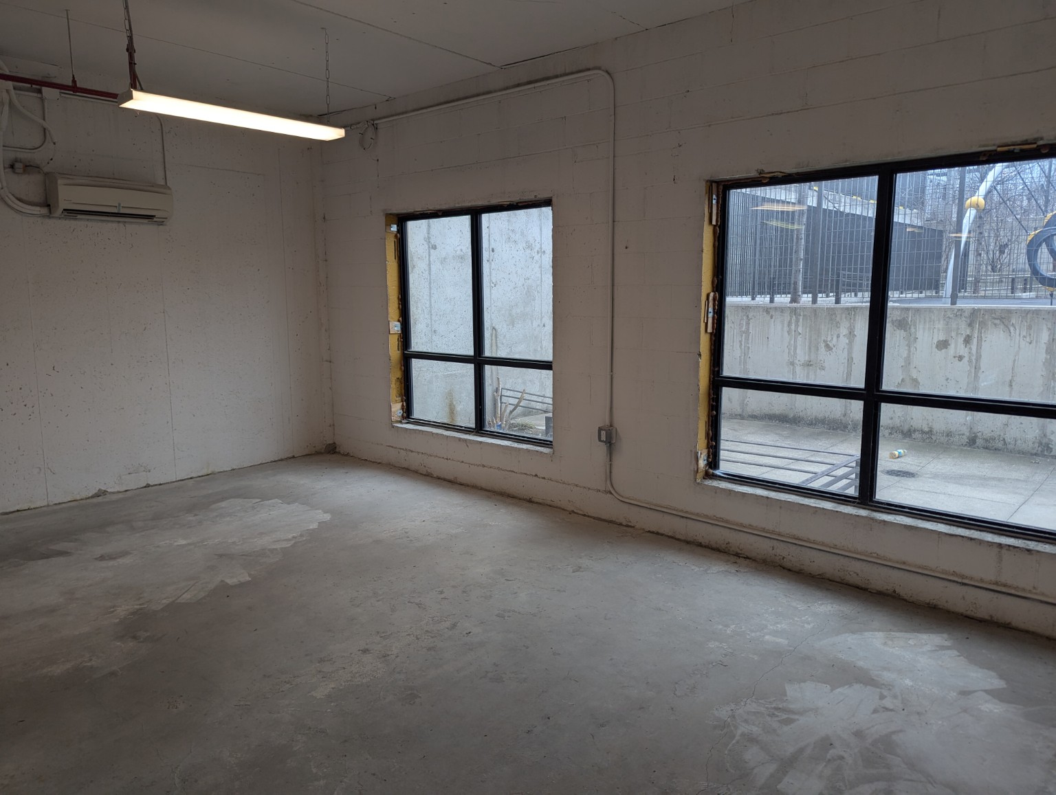 The current unfinished window-side room, with bare concrete floors, white block walls, and two large windows